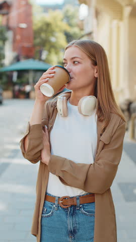 Happy Caucasian woman enjoying morning coffee hot drink and smiling. Relaxing, taking a break. Young girl teenager walking on urban city center street, drinking coffee to go. Town lifestyles outside.