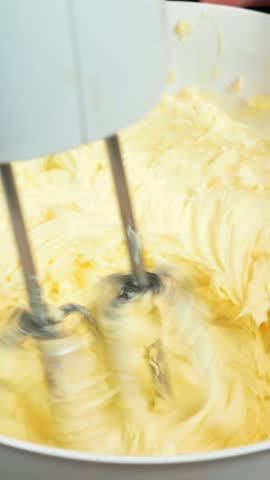Woman whisks buttercream in bowl with hand electric mixer, making cake. Cooking process. Female baker is preparing homemade cake, mixing butter and sugar. Vertical video