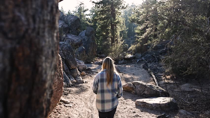 young girl wearing a light blue plaid flannel and black pants walking hiking through the forest in Big Bear California during sunset with dramatic shadows and lots of rocks and boulders STATIC SHOT