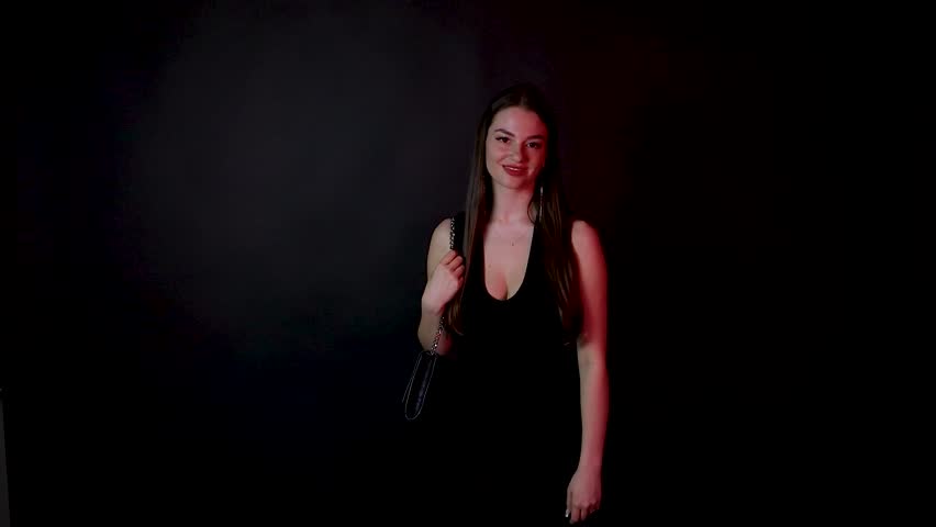 Young Caucasian female adult brunette model posing in front of a dark background wearing black dress with a purse