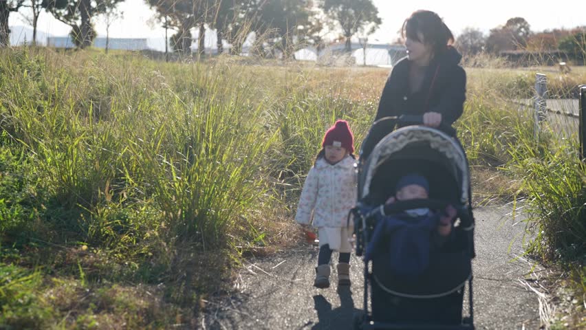 On a winter evening in December 2024, a Japanese parent and child - a 3-year-old girl, a 6-month-old boy, and a mother in her 30s - spend time in a lush park in Fukuoka City, Fukuoka Prefecture.