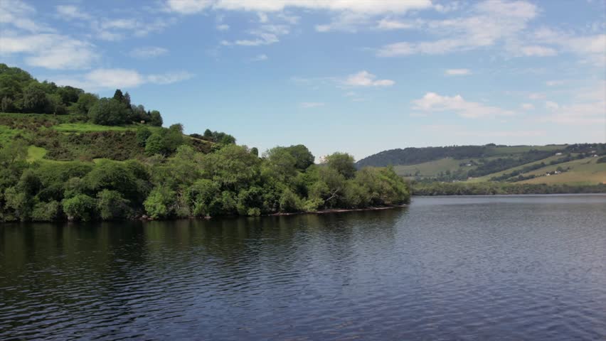 Drone footage capturing the picturesque shoreline of Loch Ness, surrounded by lush green hills and peaceful waters under a bright sky. Perfect for travel and nature themes.