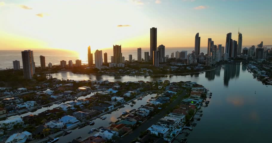 At sunrise, a drone view of Surfers Paradise reveals a golden glow as sunlight reflects on calm waters. High-rises along the coast catch soft dawn hues, with the ocean mirroring the sky and skyline.