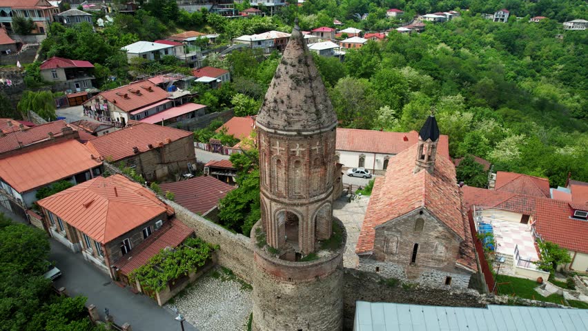 Aerial Drone Footage Showcases An Ancient Tower In Sighnaghi, Georgia, Surrounded By Lush Green Trees On A Cloudy Summer Day. The Camera Gracefully Pans, Revealing Stunning Mountain Views.
