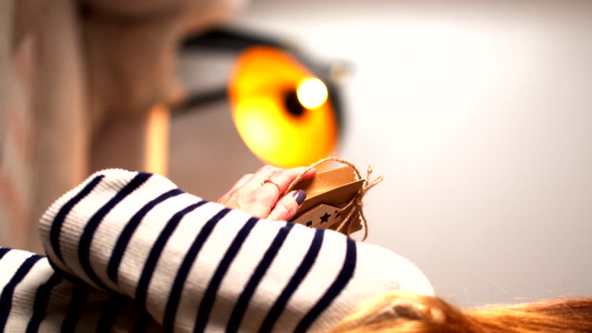 Female hand in a striped sweater holds a small rustic Christmas gift box tied with twine and decorated with festive cutouts. A warm, glowing light in the blurred background adds a cozy holiday feel.