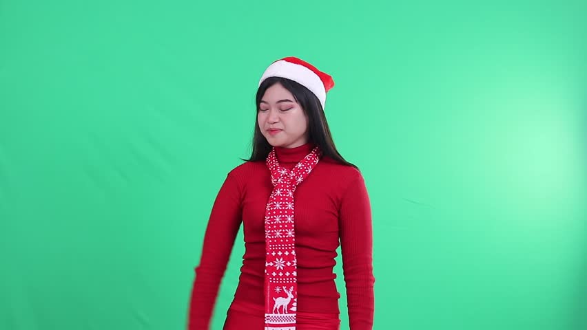 Asian woman charm wearing christmas sweater, Santa hat and scarf, shocked facing camera looking to the right while covering mouth, isolated on green background, green screen