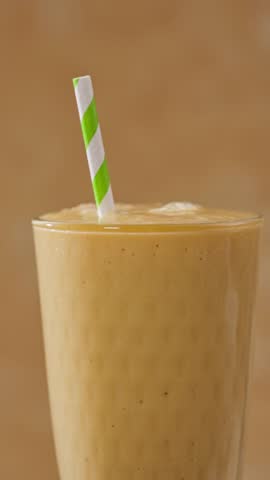 glass of fresh yellow banana mango and orange smoothie with straw turning and rotating on yellow background