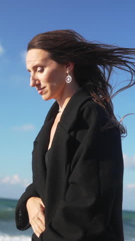 Woman Beach Coat Wind, A woman wearing a black coat stands on a beach with the wind blowing her hair, the sky and ocean in the background. Woman Beach Ocean Waves - A woman sits on a rock on a beach