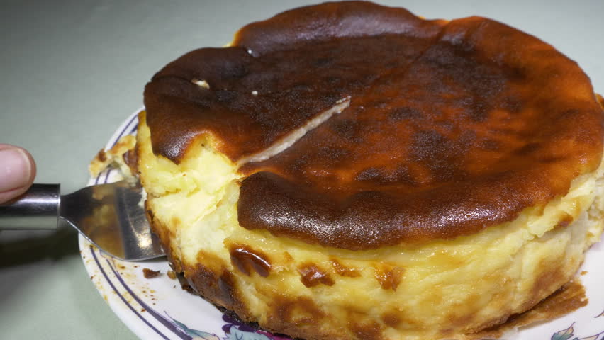 Closeup of a steel cake serving tool carefully lifting up and pulling out the first triangular slice of a whole round, soft, baked cheesecake dessert on a plate.