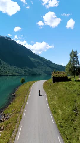 A cyclist enjoys a tranquil ride next to a stunning fjord in Norway. The sun shines brightly, illuminating the lush green mountains and picturesque landscape winding road. Lovatnet Norway