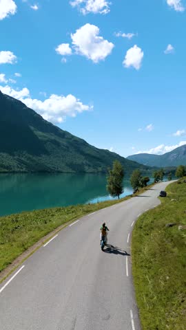 A cyclist enjoys a tranquil ride next to a stunning fjord in Norway. The sun shines brightly, illuminating the lush green mountains and picturesque landscape winding road. Lovatnet Norway