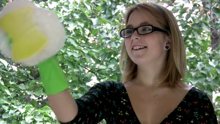 Young and pretty girl removes the foam from the window with the scraper and the smiles on the background of green leaves