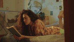 Young long-haired woman lying on stomach on bed and reading book in cozy sunlit room with plants and wall decorations, resting during day at home - Powered by Shutterstock - Get 15% off with code: PIKWIZARD15