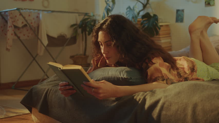 Girl reading book on bed, then putting it aside and lying face down on in cozy sunlit room decorated with indoor plants and wall art, feeling bored or sad during lonely day at home. Zoom-out shot