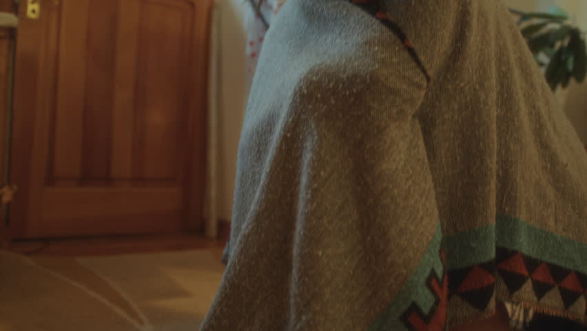 Young girl wrapped in blanket, sitting on floor in sunlit room, then walking to mirror, looking thoughtfully at her reflection and rubbing her face. Low angle view