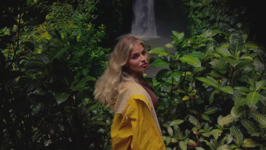 Portrait: beautiful blonde Slavic girl exploring a waterfall in Bali Indonesia. Woman in a yellow raincoat with beauty face in tropical jungle is enjoying vacation, trip. Rainy season, tropical plant
