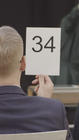 Vertical shot of auction participants sitting in audience, raising numbered cards to place bids