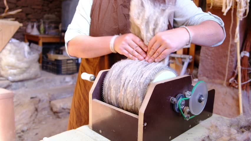 A skilled female artisan close-up demonstrates the process of carding wool using a hand-operated machine. The workshop is filled with natural wool fibers, showcasing traditional crafting techniques.