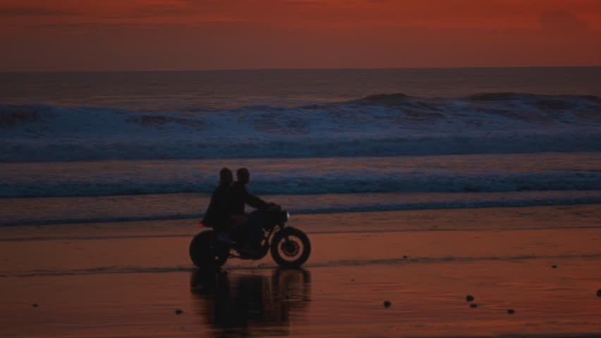 Man, woman ride vintage motorcycle or motorbike along ocean coast in Bali. Couple sits and riding on a scooter on sandy sea beach, sunset. Travelers enjoy journey on bike, love on adventure, road trip