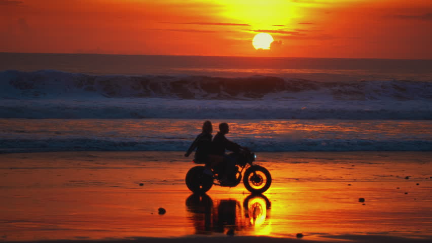 Man, woman ride vintage motorcycle or motorbike along ocean coast in Bali. Couple sits and riding on a scooter on sandy sea beach, sunset. Travelers enjoy journey on bike, love on adventure, road trip