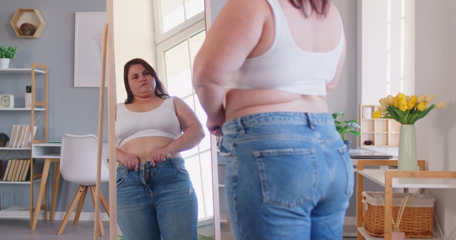 Pleasant obese woman posing in front of mirror at home, chubby overweight plus size big fat fit lady doing waist measurement with progress in weight loss after sport, dieting, looking in despair 