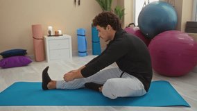 Young man stretching on yoga mat in indoor gym surrounded by exercise balls and equipment, focused on fitness routine. - Powered by Shutterstock - Get 15% off with code: PIKWIZARD15