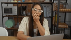 Woman yawning in a cozy home office with laptop, surrounded by shelves and kitchenware, expressing fatigue during work at home. - Powered by Shutterstock - Get 15% off with code: PIKWIZARD15