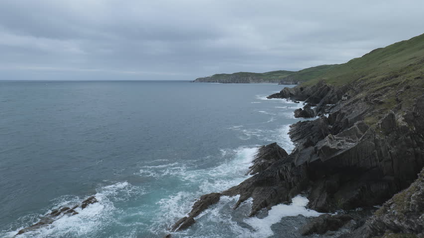 Rocky coastline with waves and green hills under a cloudy sky.