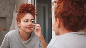 Young woman using eyelash curler in front of bathroom mirror during morning routine - Powered by Shutterstock - Get 15% off with code: PIKWIZARD15
