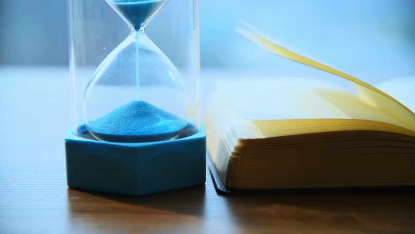 Sand running through the hourglass on wooden table with book