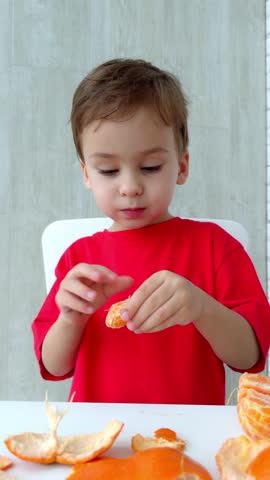 Child enjoying an orange snack indoors. Young child dressed in red savors a fresh orange while seated at a white table in a bright room. Vertical video