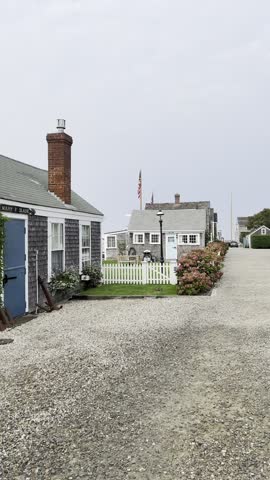 A charming Nantucket street scene featuring quaint houses, a white picket fence, hydrangeas, and a view of the American flag, framed by a gravel road and a lamp post in the peaceful neighborhood.