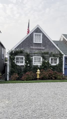A charming Nantucket house with a flag, the word “Constitution” on the front, surrounded by blooming flowers and a fire hydrant, set against a serene sky and gravel pathway.