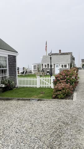 A charming Nantucket street scene featuring a white picket fence, colorful hydrangeas, a blue door cottage with an American flag, and a curious rabbit in the grassy yard by the gravel path.