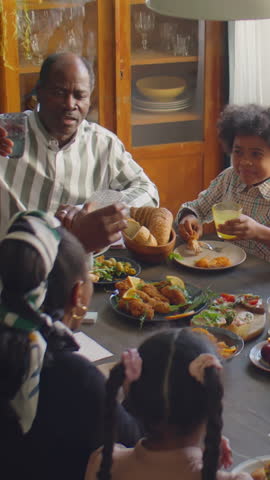 Vertical shot of African American family gathered around dinner table raising glasses in toast during holiday celebration at home