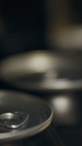 A close-up shows several beverage cans resting on a dark surface, highlighting the silver tops and pull tabs. The composition emphasizes the metallic textures and reflections visible on the cans