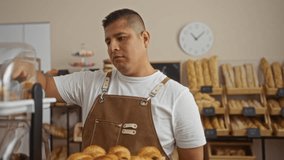 Young hispanic man working in a bakery shop arranging pastries indoors wearing an apron with pastry shelves and bread loaves in the background - Powered by Shutterstock - Get 15% off with code: PIKWIZARD15