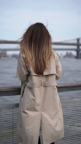 A woman in a stylish trench coat gazes at a distant city bridge by the shimmering water in a calm urban setting. Stylish woman wearing trench coat stands by water gazing beautiful city bridge
