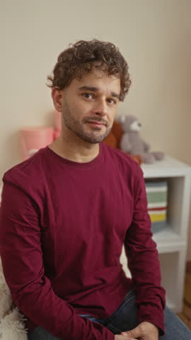Young man sitting in a cozy bedroom with a cradle, smiling warmly, dressed in a maroon shirt, providing a relaxed and homey atmosphere with soft toy accents.