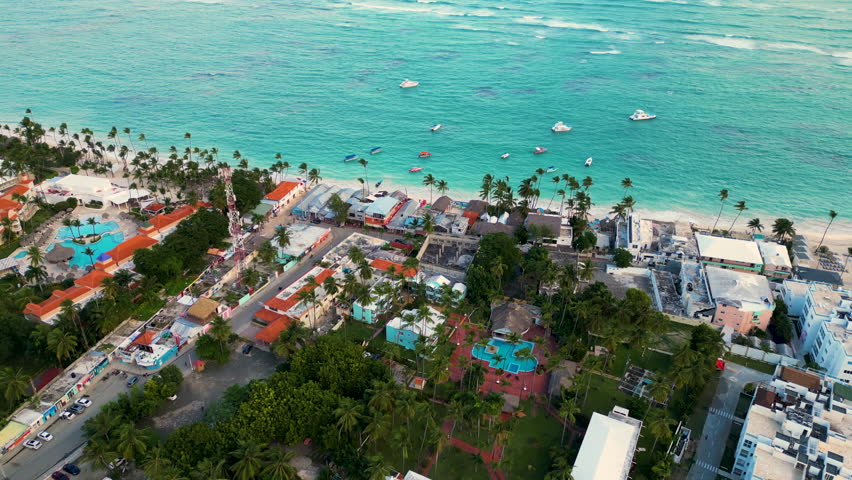 Aerial view of a vibrant beachfront area in Punta Cana, Dominican Republic, highlighting colorful buildings with red-tiled roofs surrounded by lush palm trees.