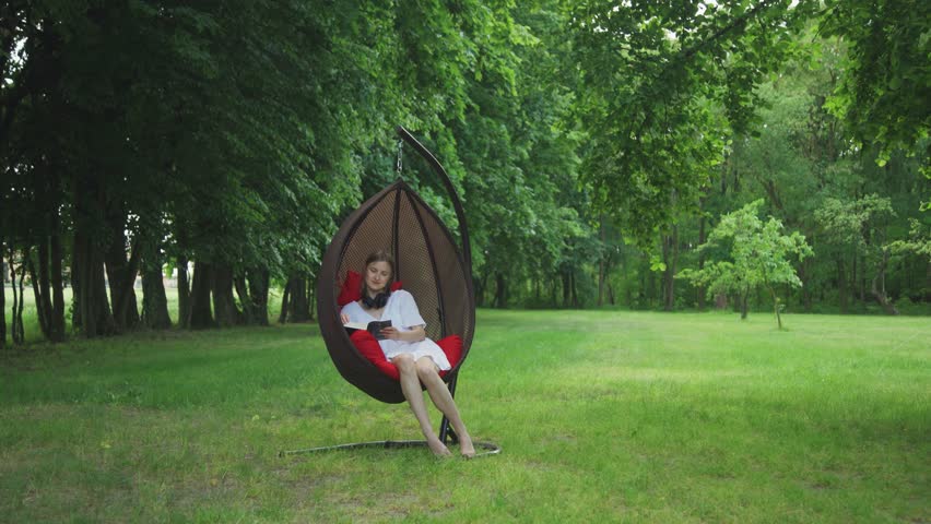 Vacation away from hustle and bustle of city. A woman in a hanging chair in nature reads a book, enjoying singing of birds. Concept of no stress, enjoying life. Breathe deeply in fresh air, calm down.