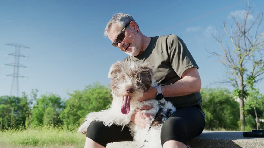 A man sits on a bench in a park, happily playing with his fluffy dog. The sun shines brightly while they share a joyful interaction amidst green surroundings.