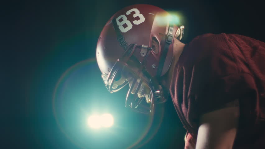 Portrait of an American football player at night at stadium lights with a helmet and protective gear uniform standing on one knee on the field holding the ball. Cinematic slow motion side view. 