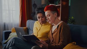 Relaxed women with laptop and tablet communicate sitting on sofa in living room. Happy lesbian couple engages in online shopping using modern devices - Powered by Shutterstock - Get 15% off with code: PIKWIZARD15