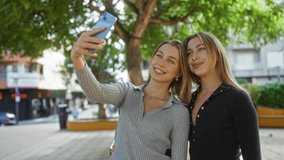Two smiling women take a selfie in an urban park setting, showcasing friendship, happiness, and modern lifestyle with trees and buildings in the background. - Powered by Shutterstock - Get 15% off with code: PIKWIZARD15
