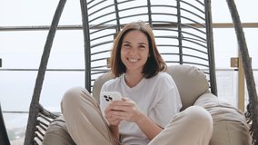 A woman sitting on a hanging chair on the balcony, relaxing and using her smartphone. - Powered by Shutterstock - Get 15% off with code: PIKWIZARD15
