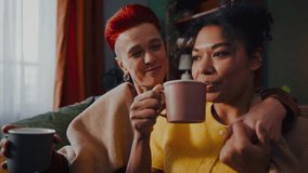 Relaxed lesbian couple holds cups enjoying hot beverages sitting on sofa in living room. Women covered with plaid enjoy romantic atmosphere at home - Powered by Shutterstock - Get 15% off with code: PIKWIZARD15