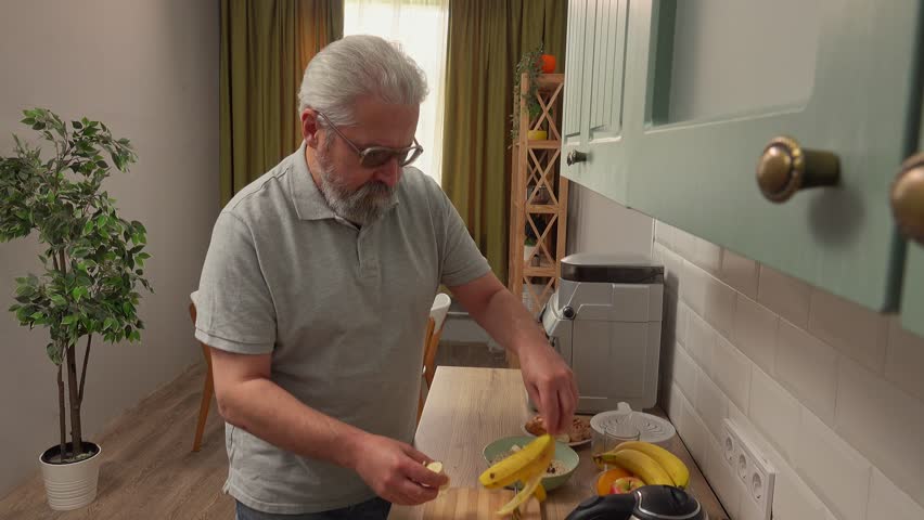 Elder grey haired man in the morning at the kitchen, prepares healthy breakfast, cuts banana and adds in bowl with oats, positive expression. Daily routine lifestyle concept