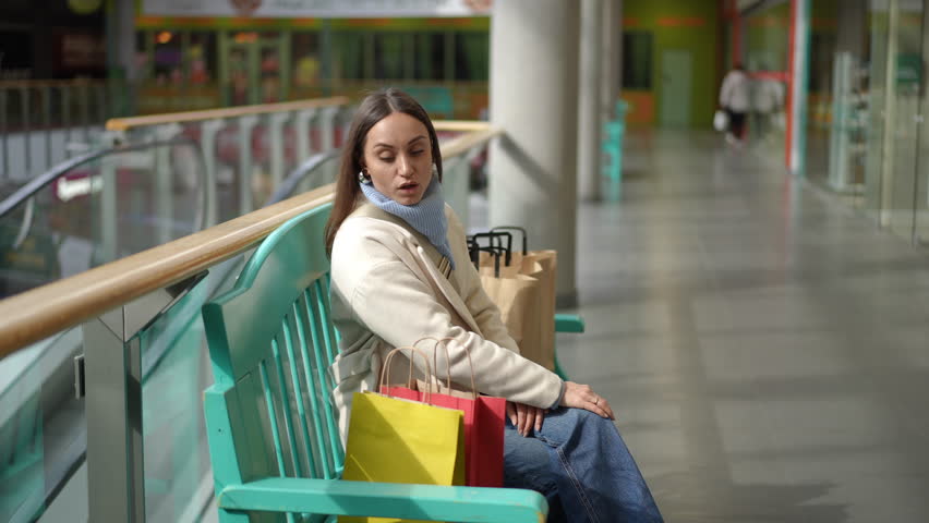 A woman sits on a turquoise bench in an outlet mall, visibly tired after a day of Black Friday shopping. Surrounded by colorful shopping bags, she leans back, taking a moment to rest. The spacious