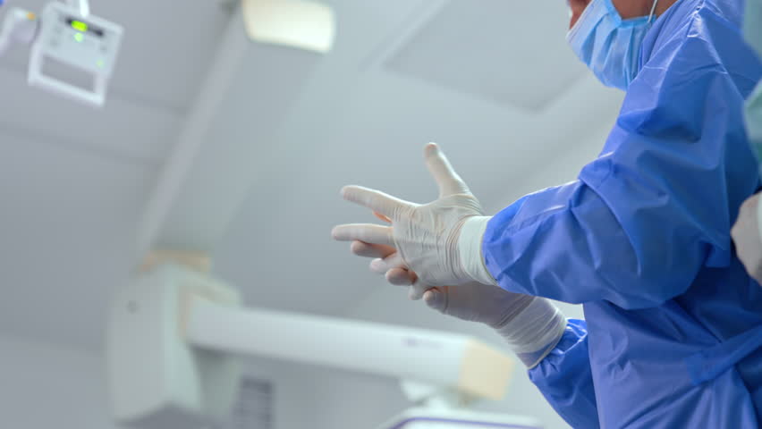 Unrecognized surgeon in blue robe putting on latex gloves. Assistant tying the coat. Low angle view. Preparation for operation. - Powered by Shutterstock - Get 15% off with code: PIKWIZARD15